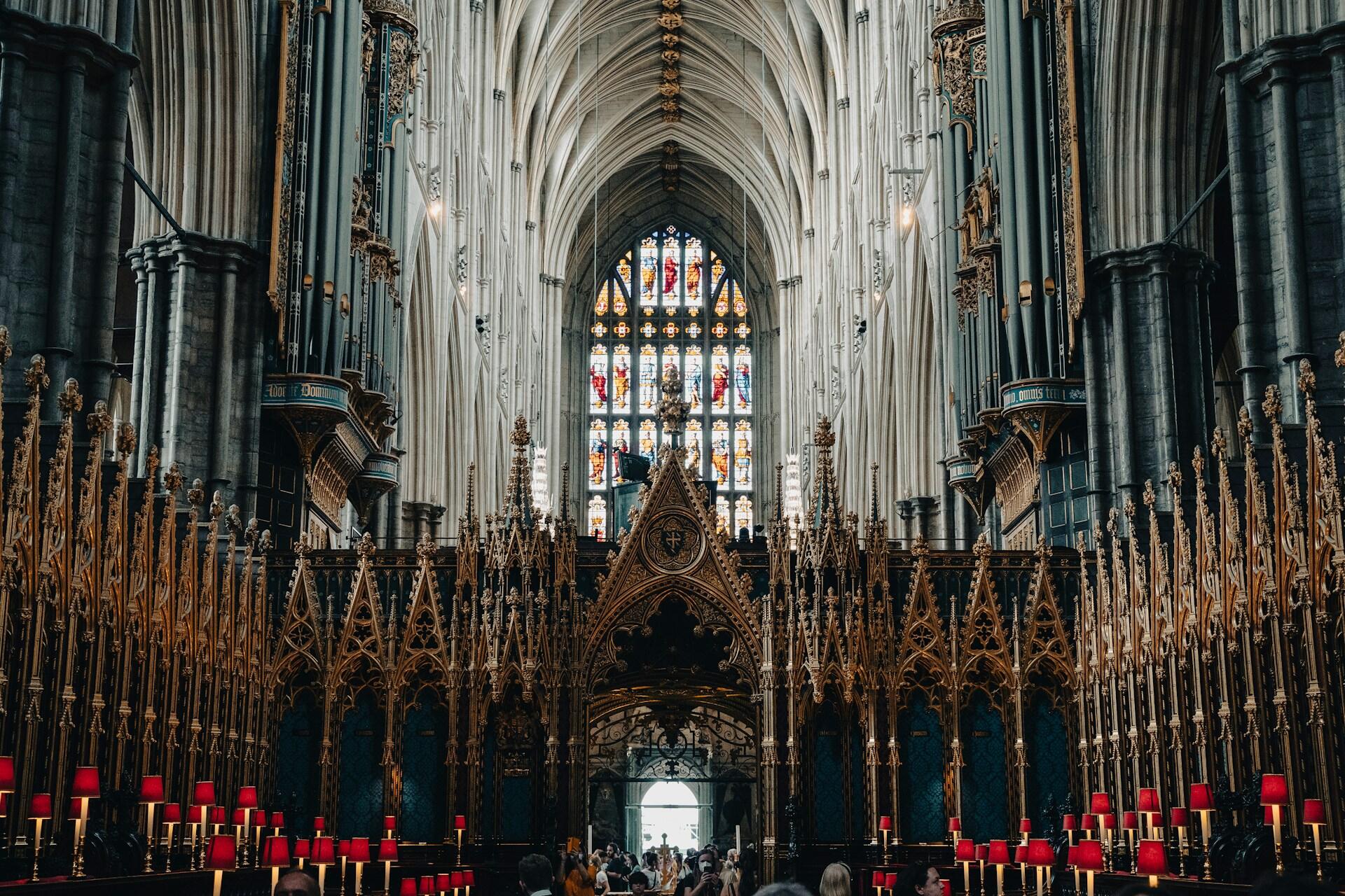 Interior of a grand cathedral featuring tall arches, intricate woodwork, stained glass windows, and rows of candles with red lamps.