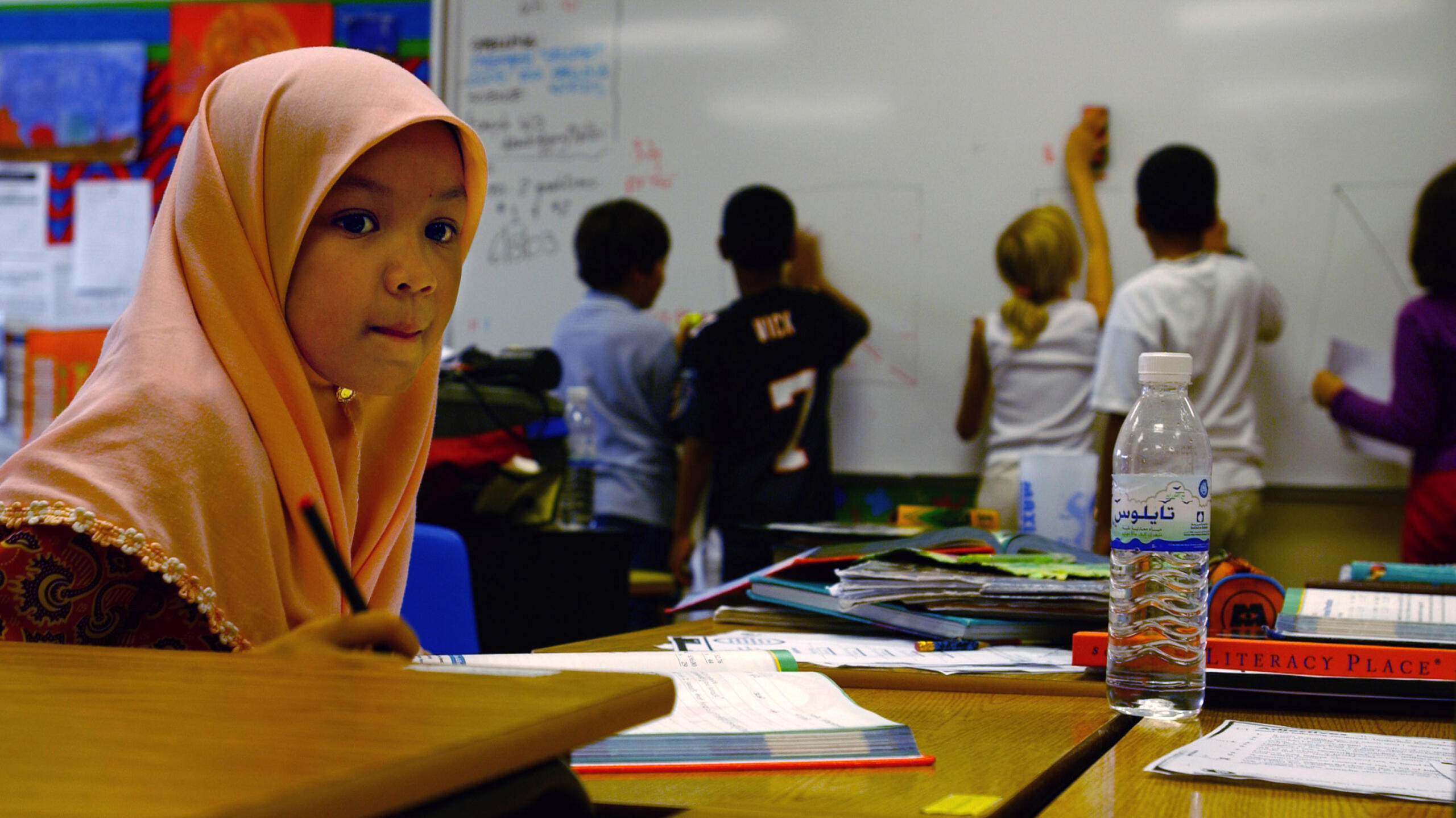 a young child with a headscarf sitting in a classroom working, with children writing on a whiteboard in the background