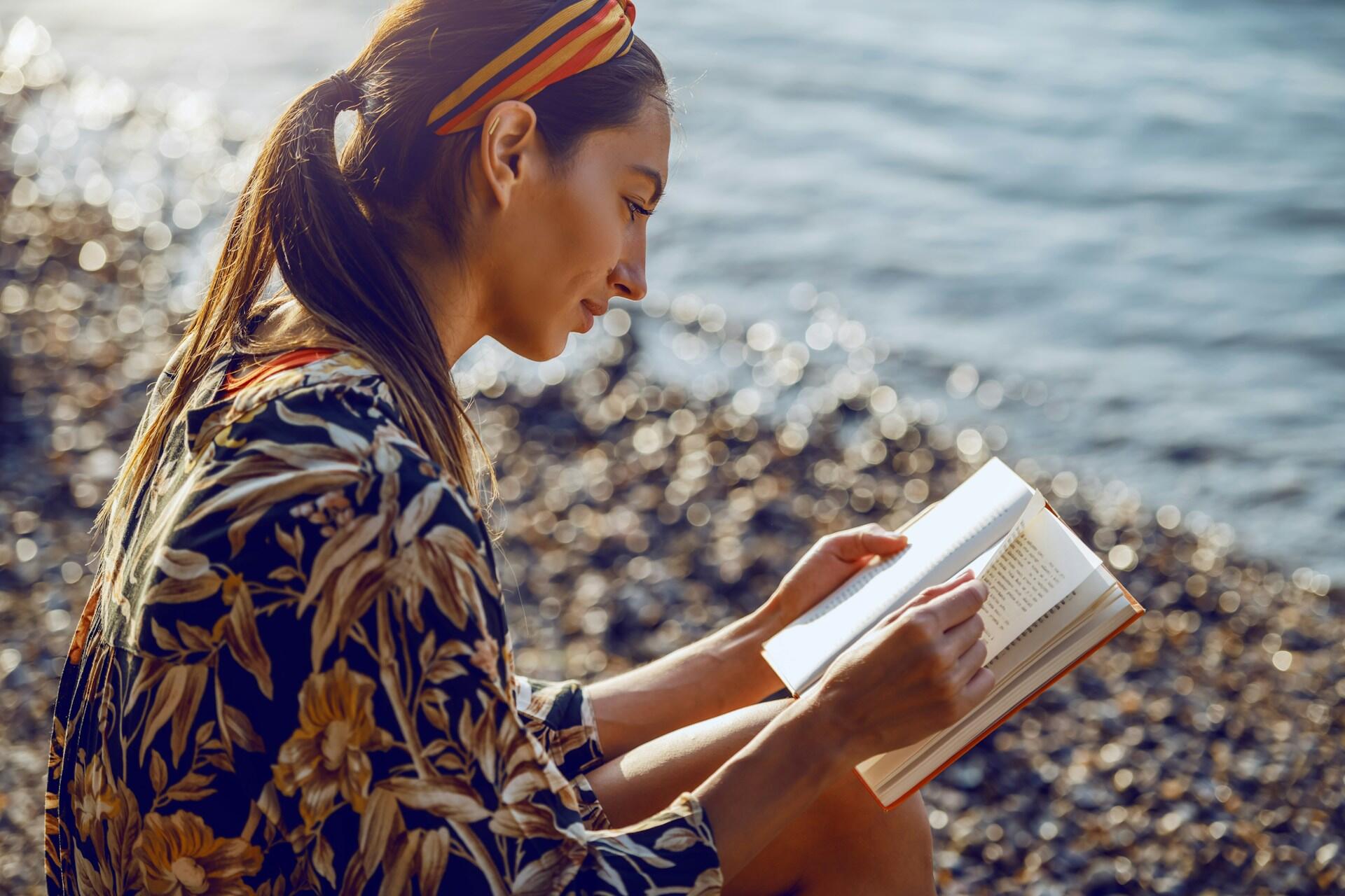 A woman with her hair pulled back, wearing a headband, sits on a beach, reading a book. The mood is tranquil.