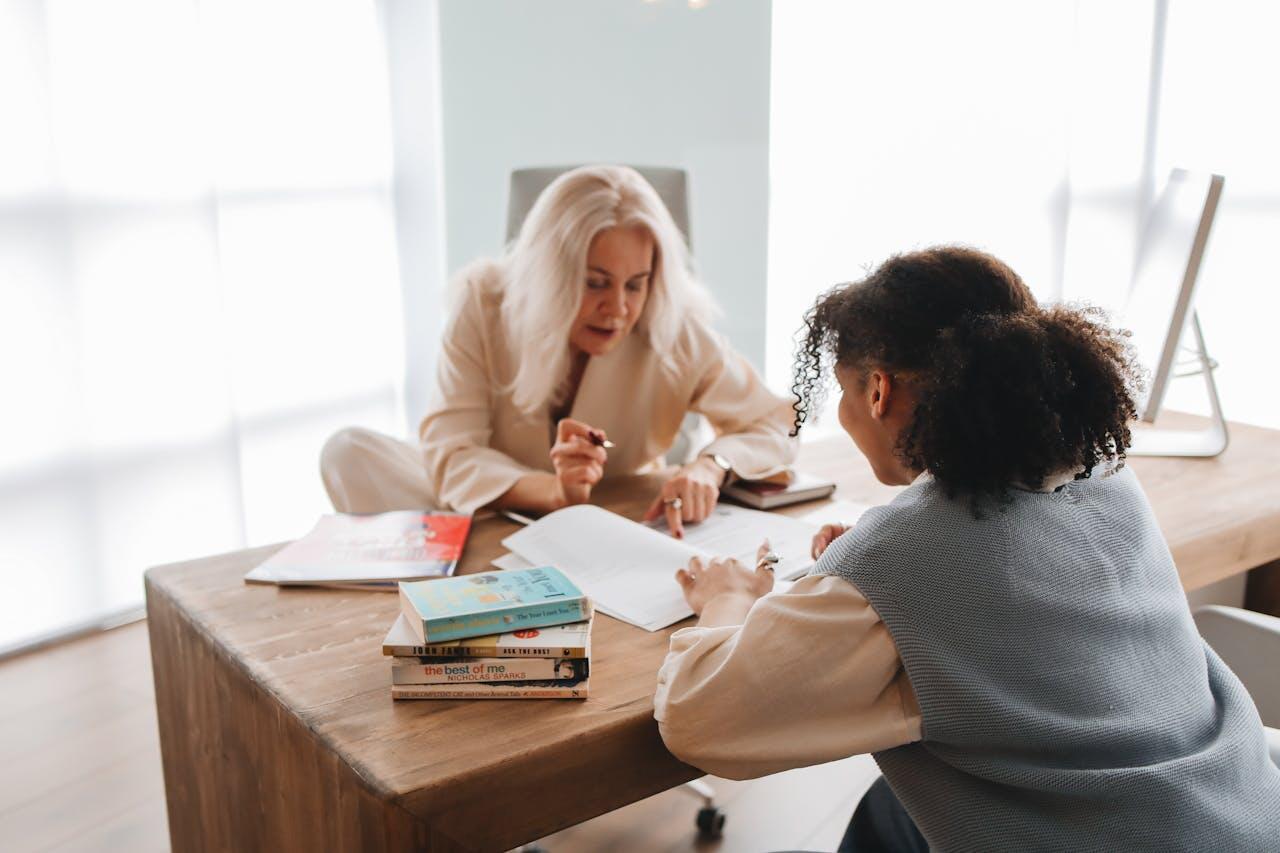 Two women engage in a discussion at a wooden table filled with books and papers, surrounded by natural light.
