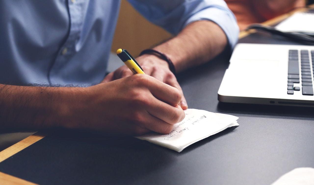 person writing in a notebook beside an open laptop