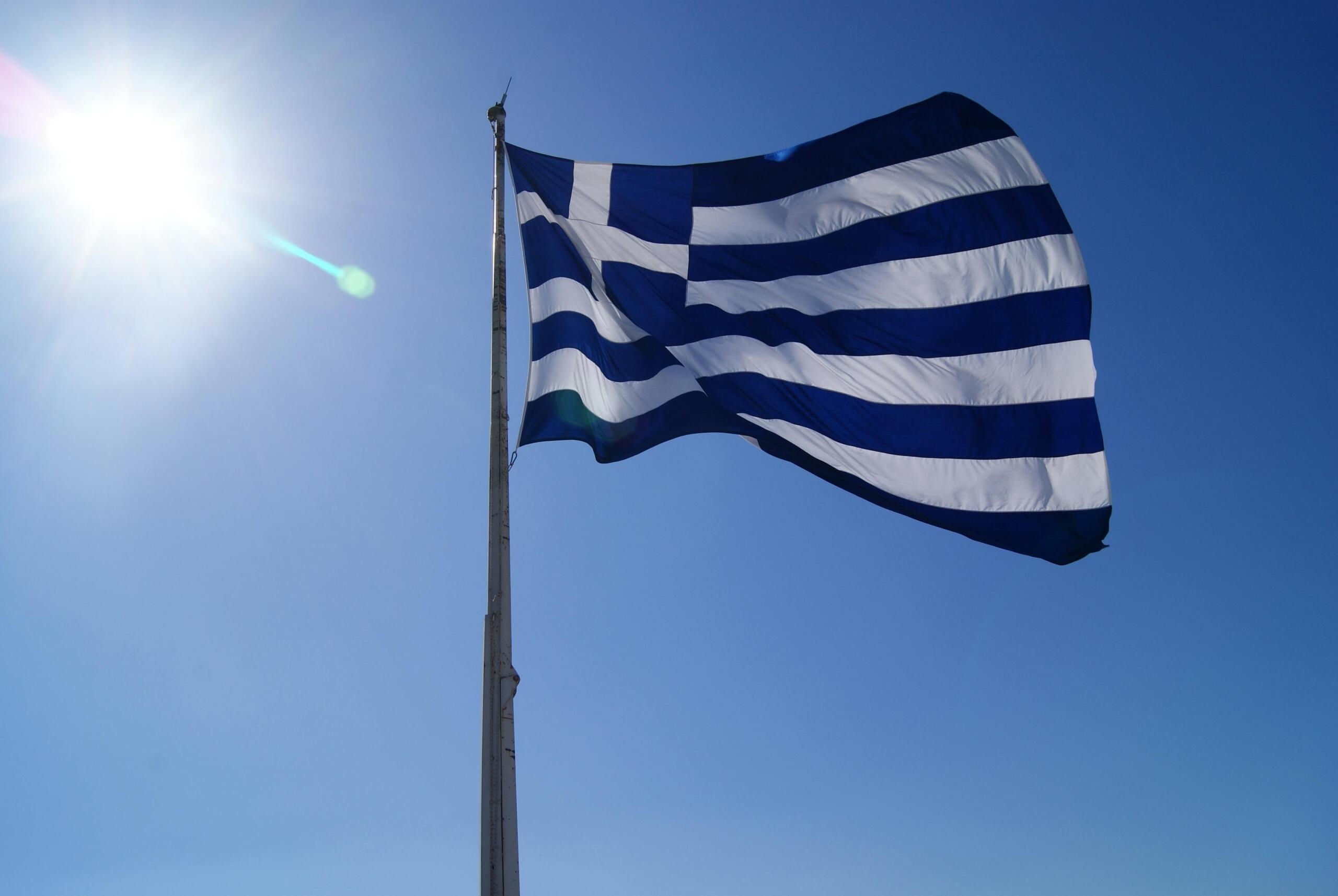 Greek flag fluttering in the wind against a bright blue sky. Ask ChatGPT