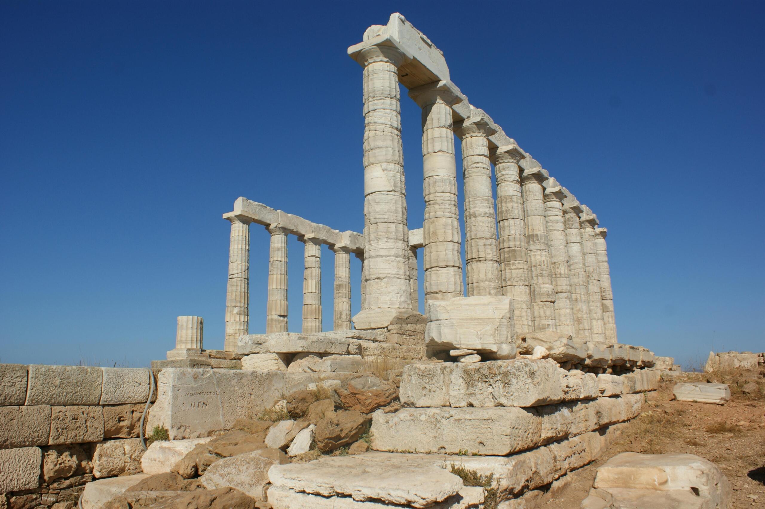 Ancient Greek ruins featuring tall marble columns and stone foundations set against a clear blue sky.