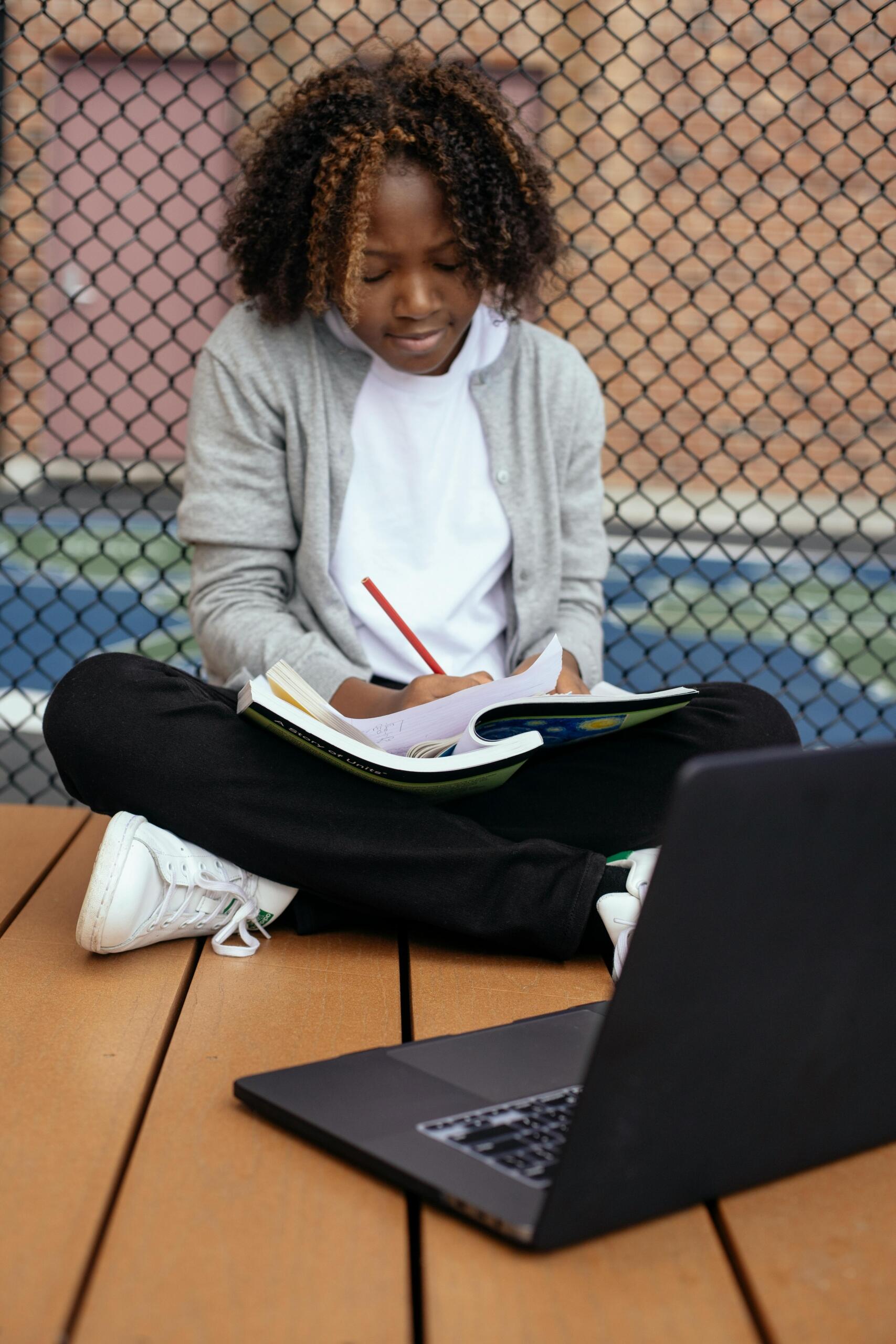 Girl sitting cross-legged on the floor, writing in a notebook with an open laptop in front of her.
