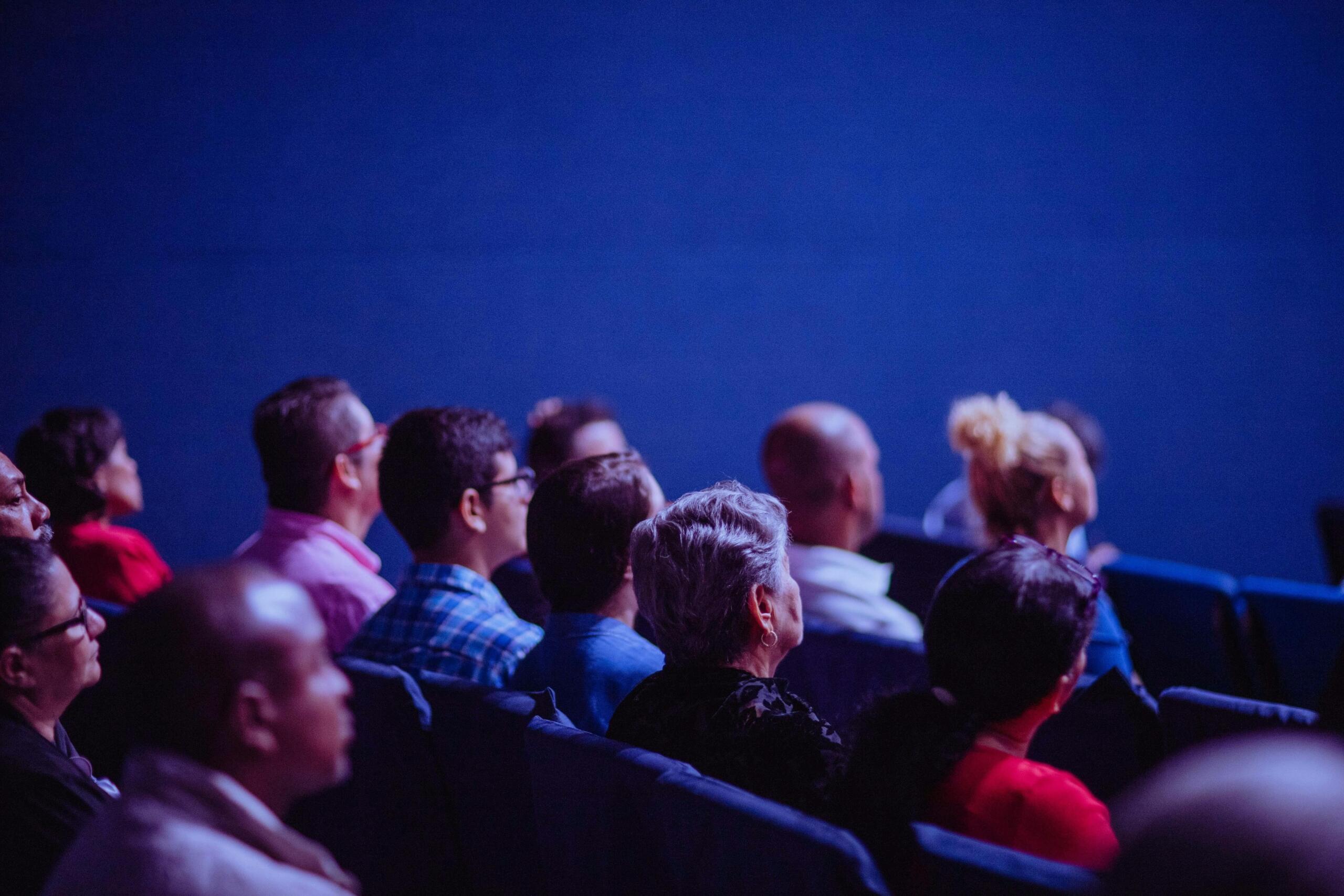 Audience sitting in a dark movie theater, watching a film on a large screen with rows of seats visible.