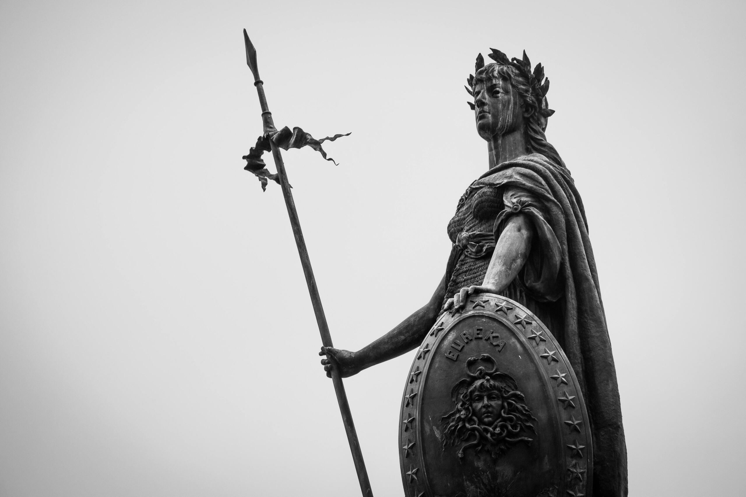 Sculpture of a Greek god holding a weapon and shield, with detailed muscular features, set against a black and white backdrop.