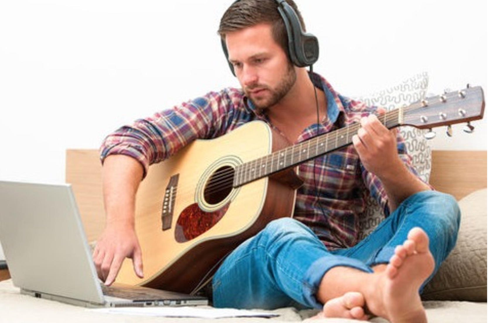 A man with headphones and a guitar sitting in front of his laptop.