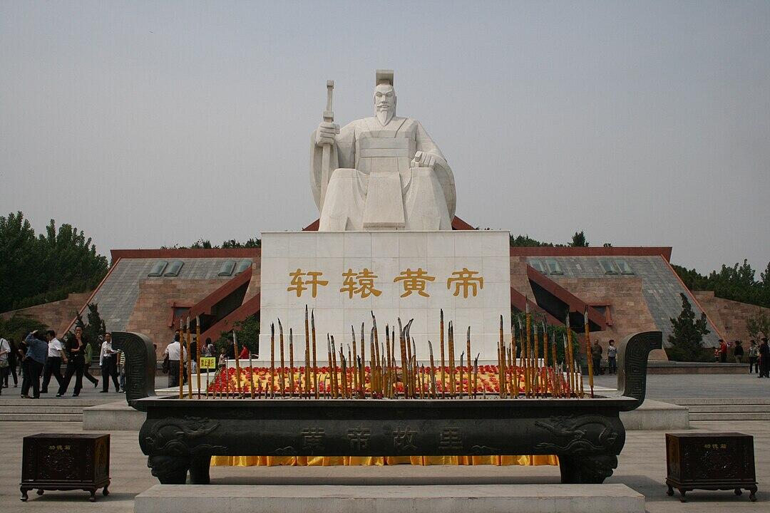 A large white statue of a seated figure with a staff, situated in front of a stone backdrop, surrounded by visitors and offerings.