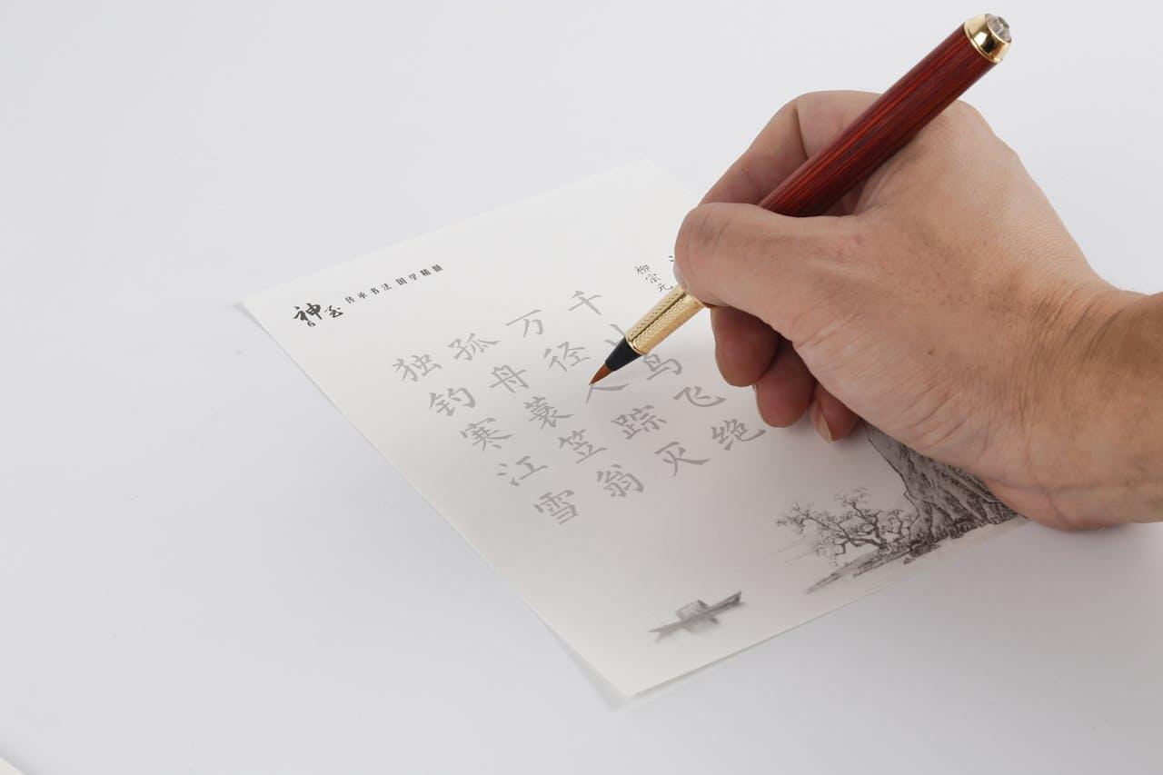 A hand writes Chinese characters with a fountain pen on a decorative letterhead featuring a boat and tree illustration.