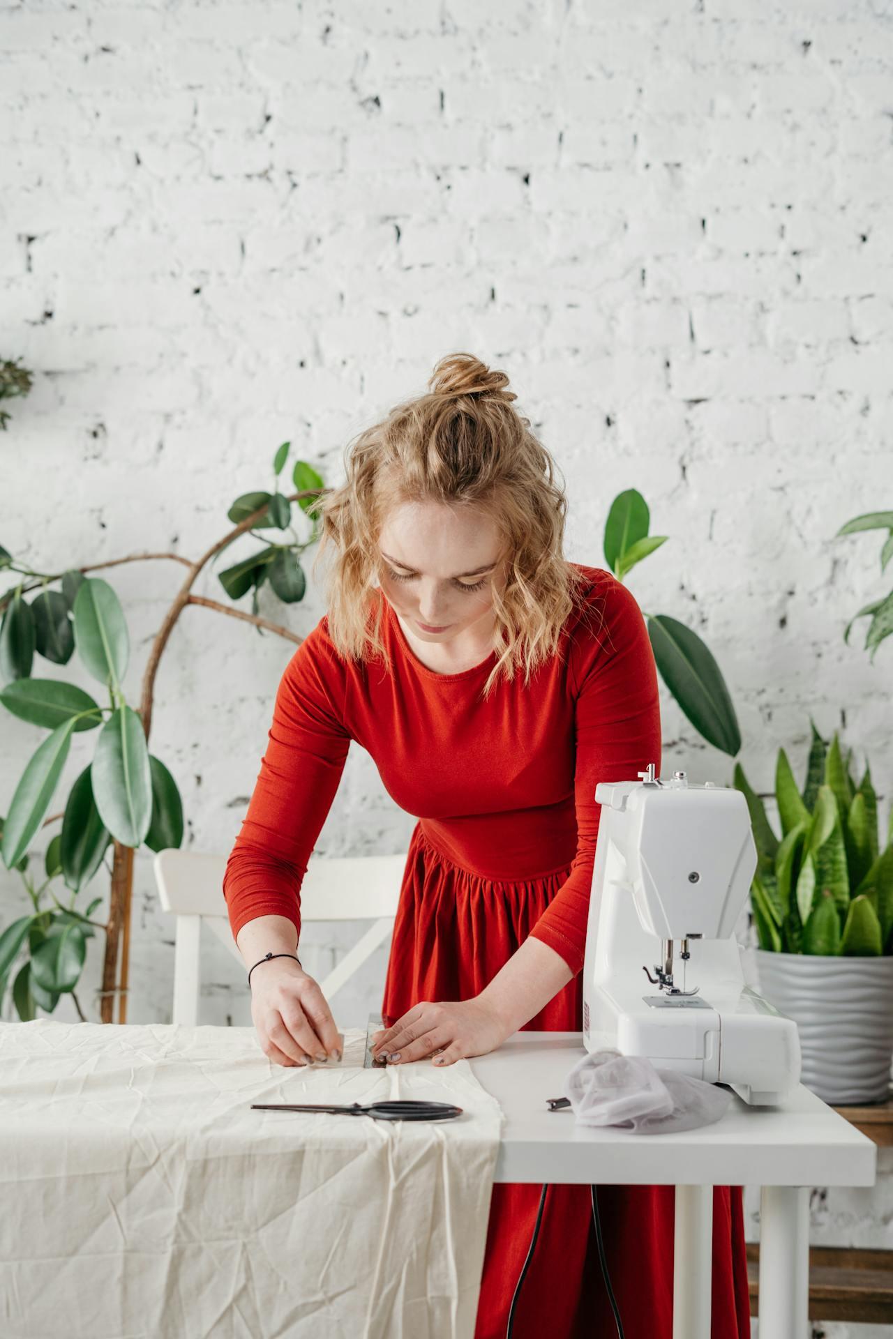 A person in a red dress is sewing fabric on a white table, surrounded by plants and a sewing machine, in a bright, minimalistic space.