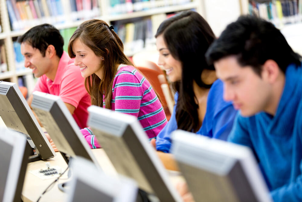 Four smiling students sitting in a row in a campus library, in front of computer monitors.