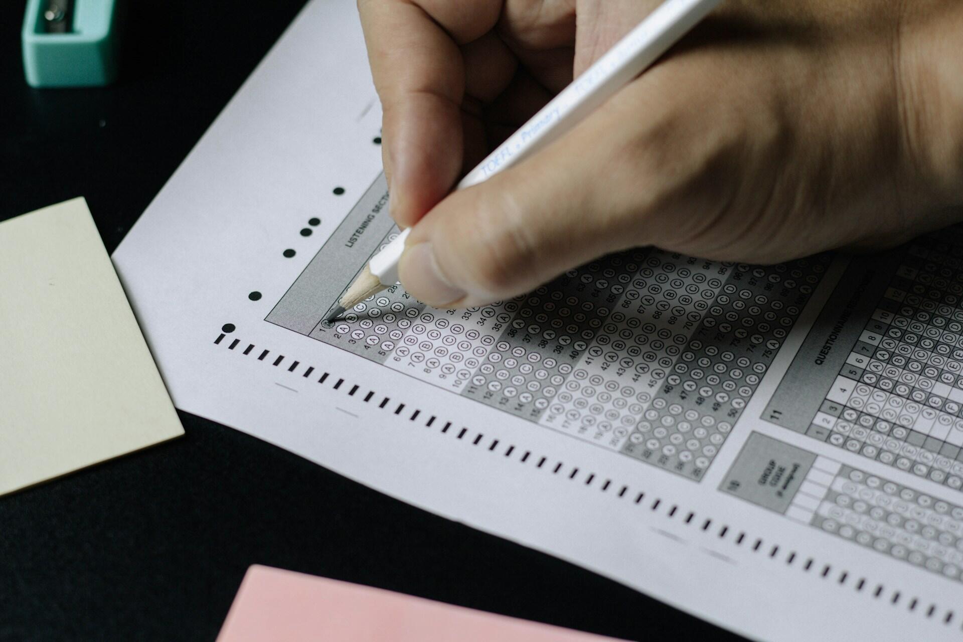 A hand holding a pencil is marking answers on a printed answer sheet, alongside colored paper notes and a stapler.