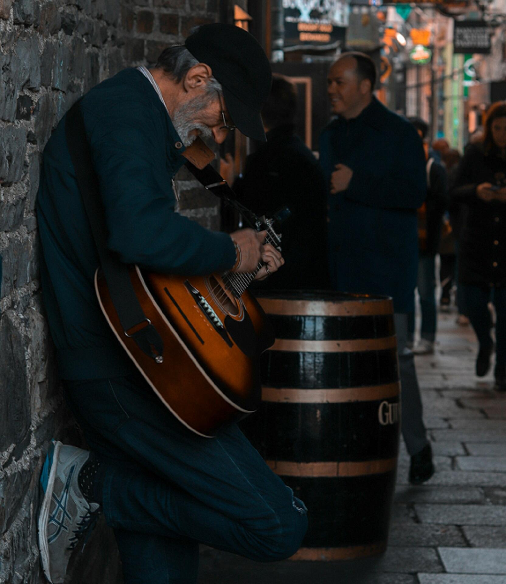 A man playing guitar leans against a stone wall.