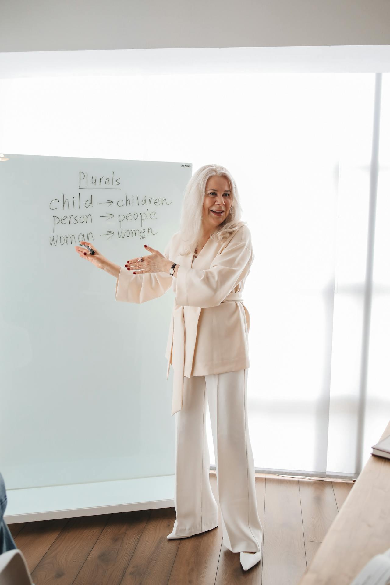 A person stands in front of a whiteboard, teaching English plural forms: child to children, person to people, woman to women.
