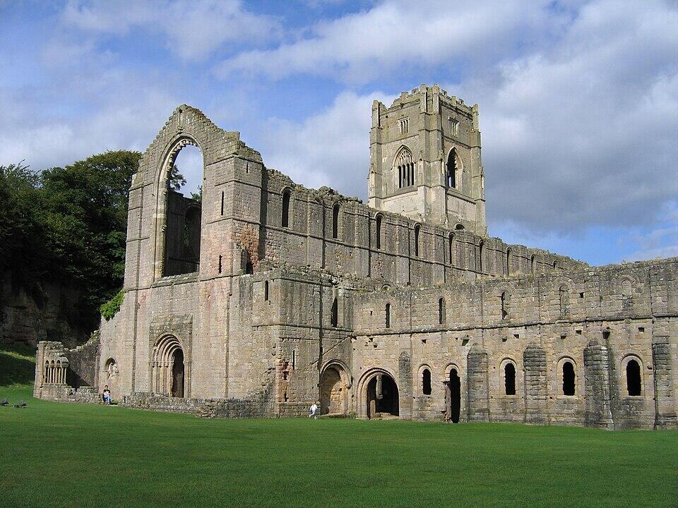 Historic ruins of a stone abbey with a tall tower, set against a blue sky with clouds, surrounded by green grass and trees.