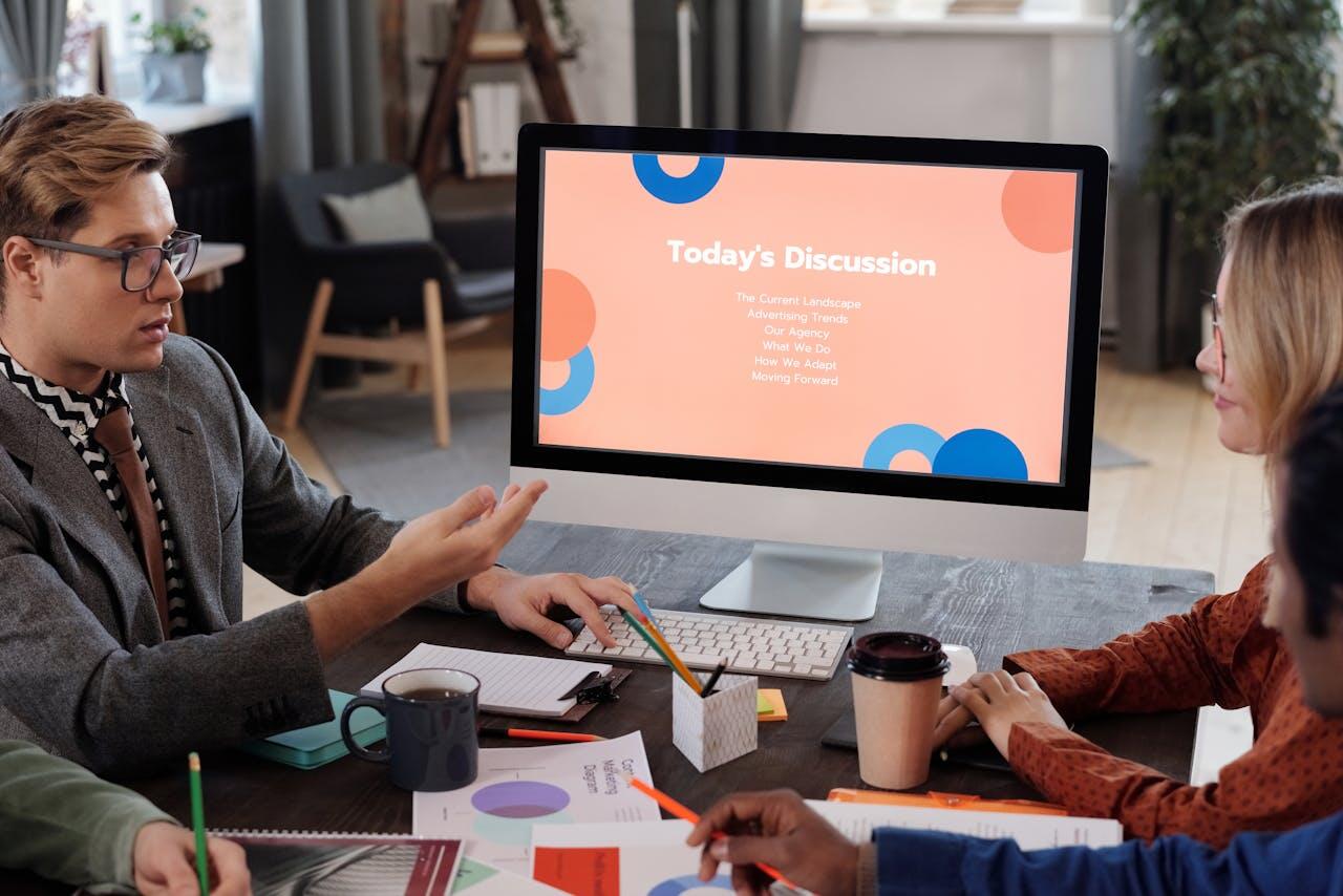 A group discusses various topics around a desk: presentations on a computer screen, notes, and coffee cups illustrate a collaborative meeting.