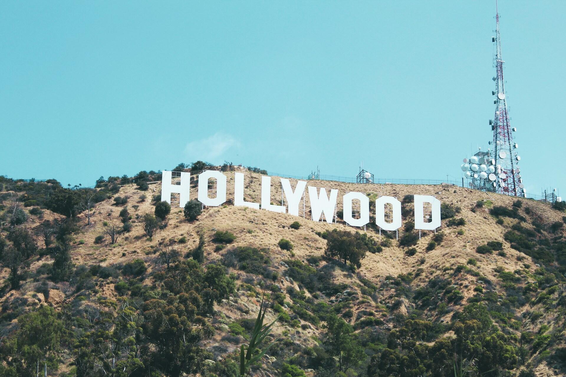 The iconic Hollywood sign stands on a hillside, with a clear blue sky and communication tower in the background.