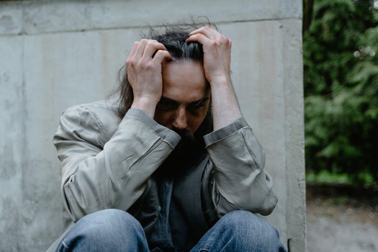 A person sits against a concrete wall, cradling their head in their hands, expressing feelings of distress or overwhelm.