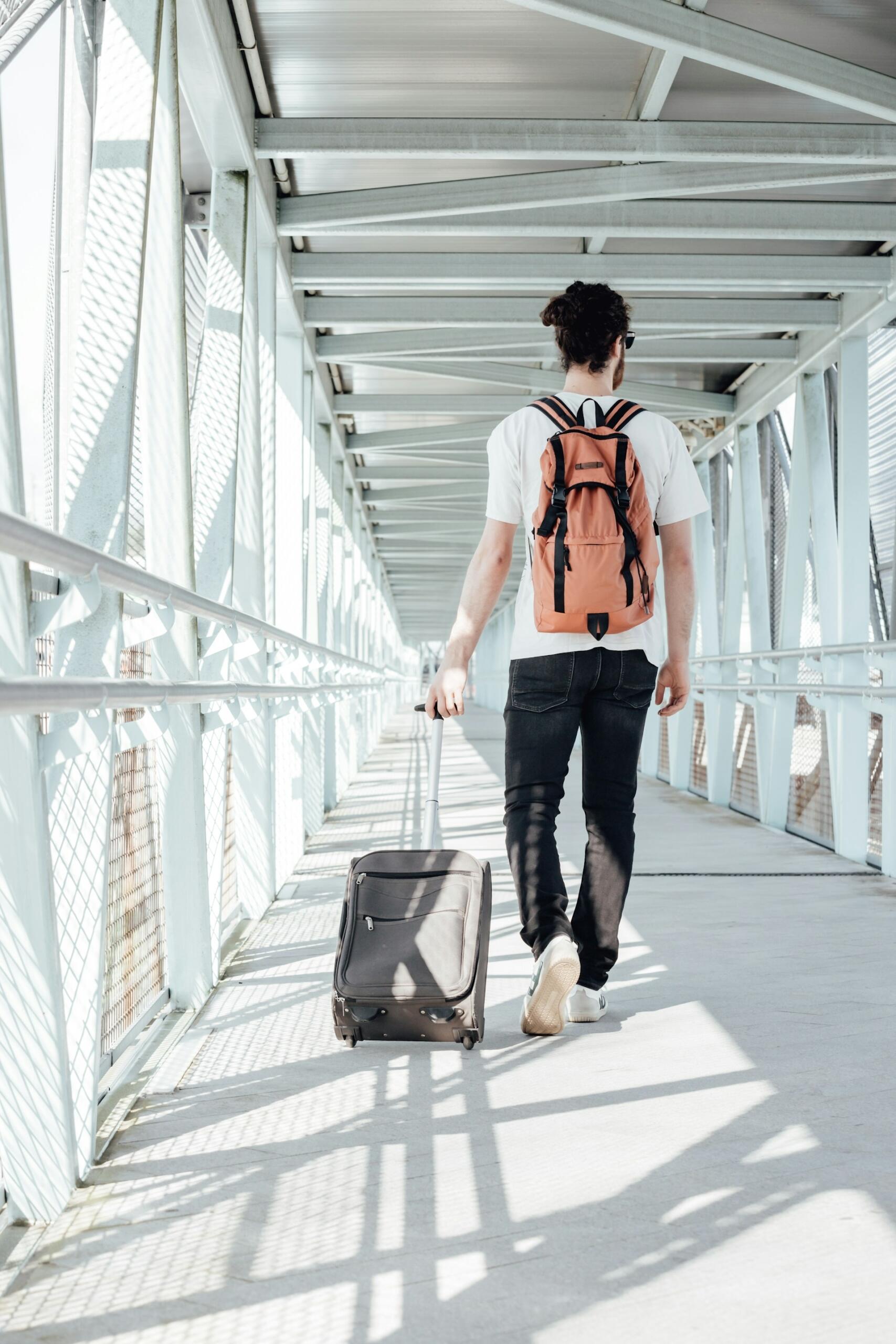 A man with an orange backpack pulling a suitcase in an airport causeway.