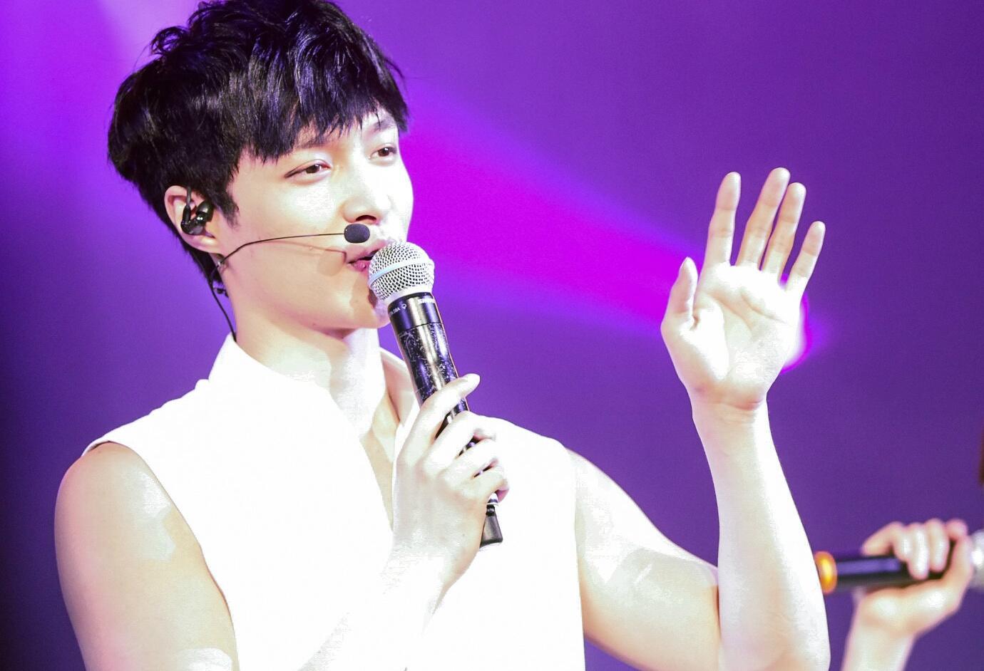 A Chinese pop artist performing in a white dress and singing with a black microphone and purple stage lights.