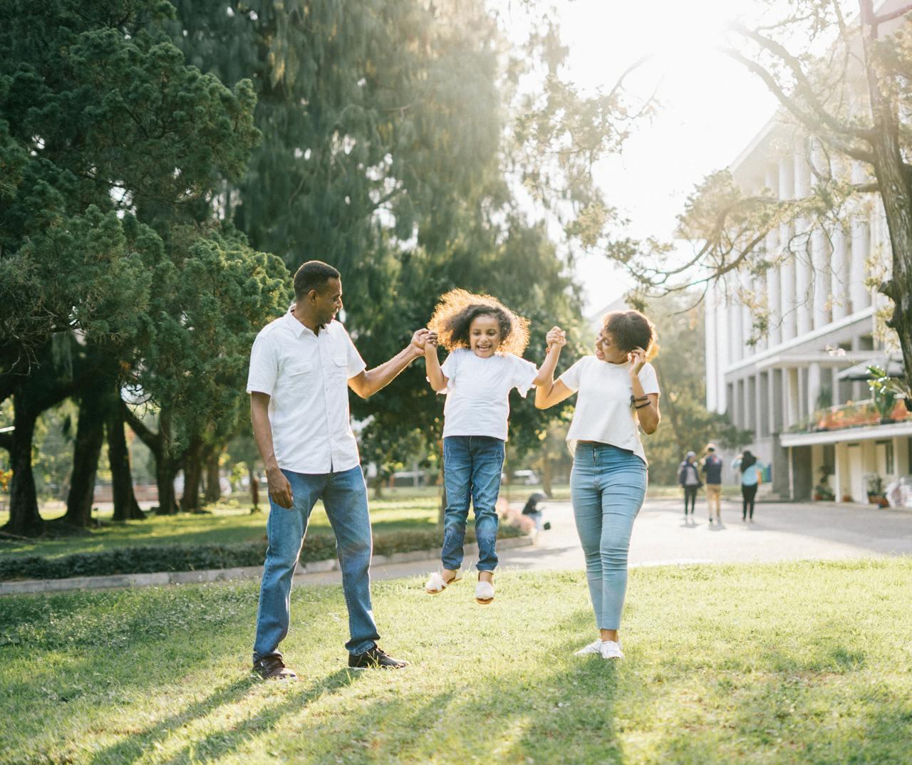 A family enjoys a sunny day outdoors, with a child being joyfully lifted between two adults in a lush green park.