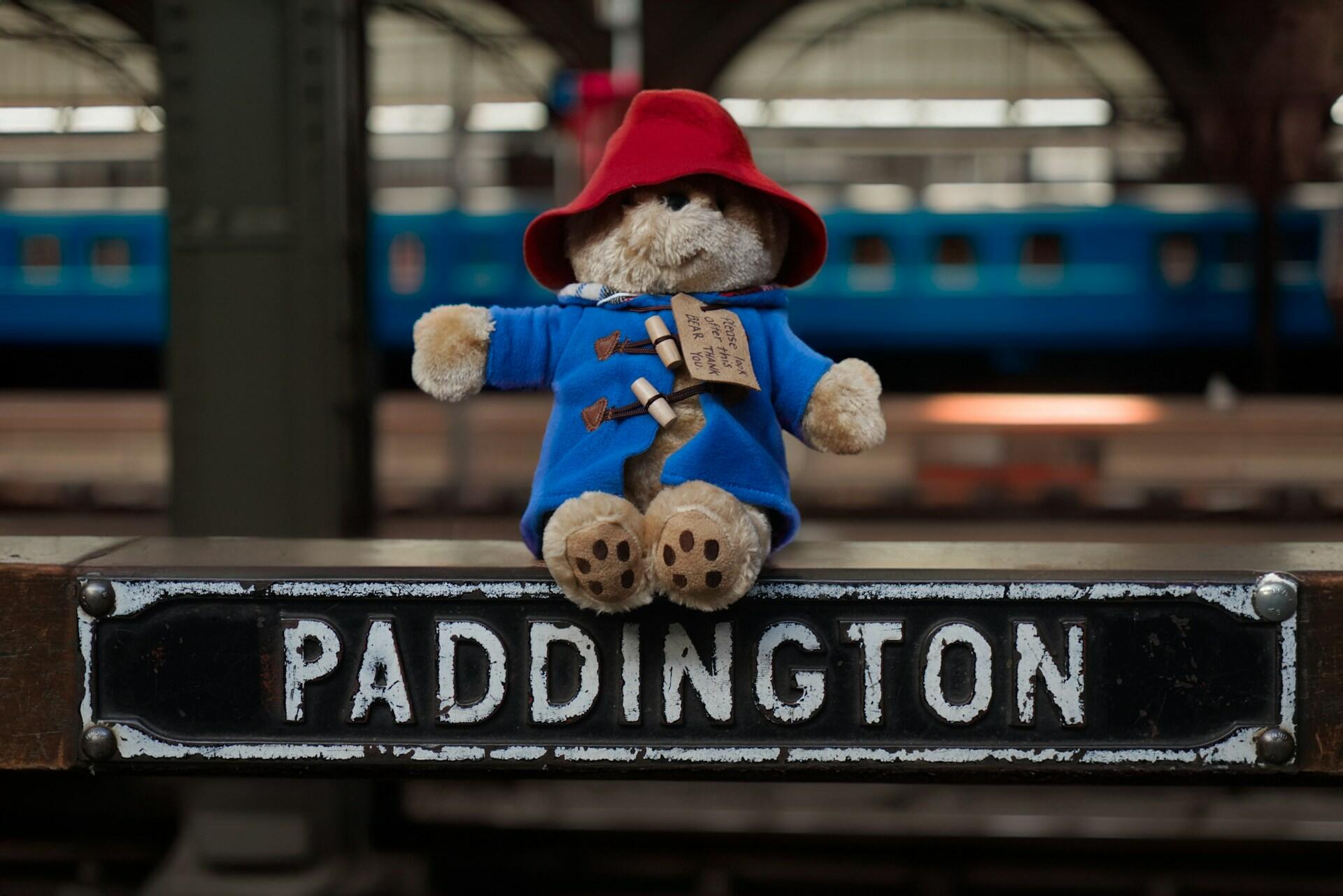 A plush bear in a blue coat and red hat sits on a "PADDINGTON" station sign, with trains blurred in the background.