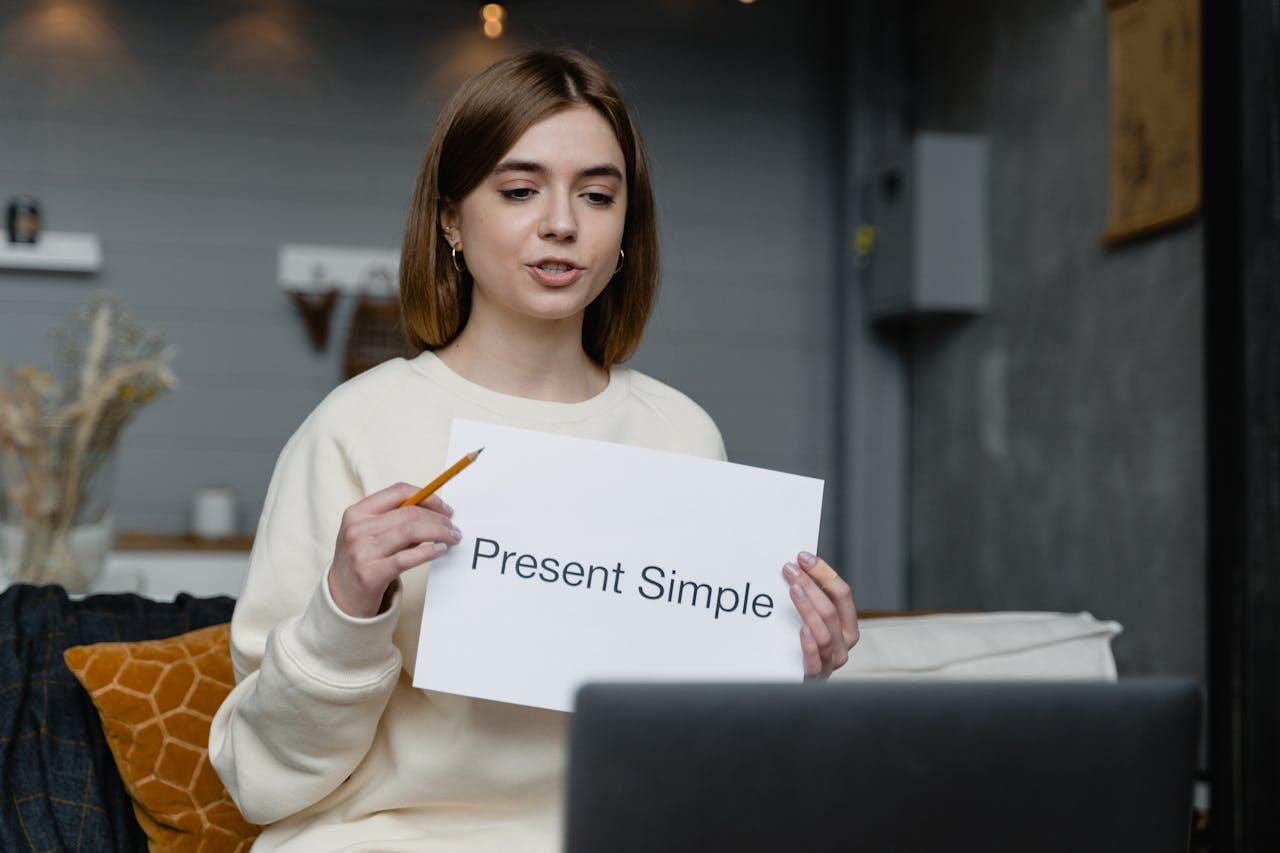 A person in a cozy room holds a sign reading "Present Simple" while sitting in front of a laptop, ready for a lesson or presentation.