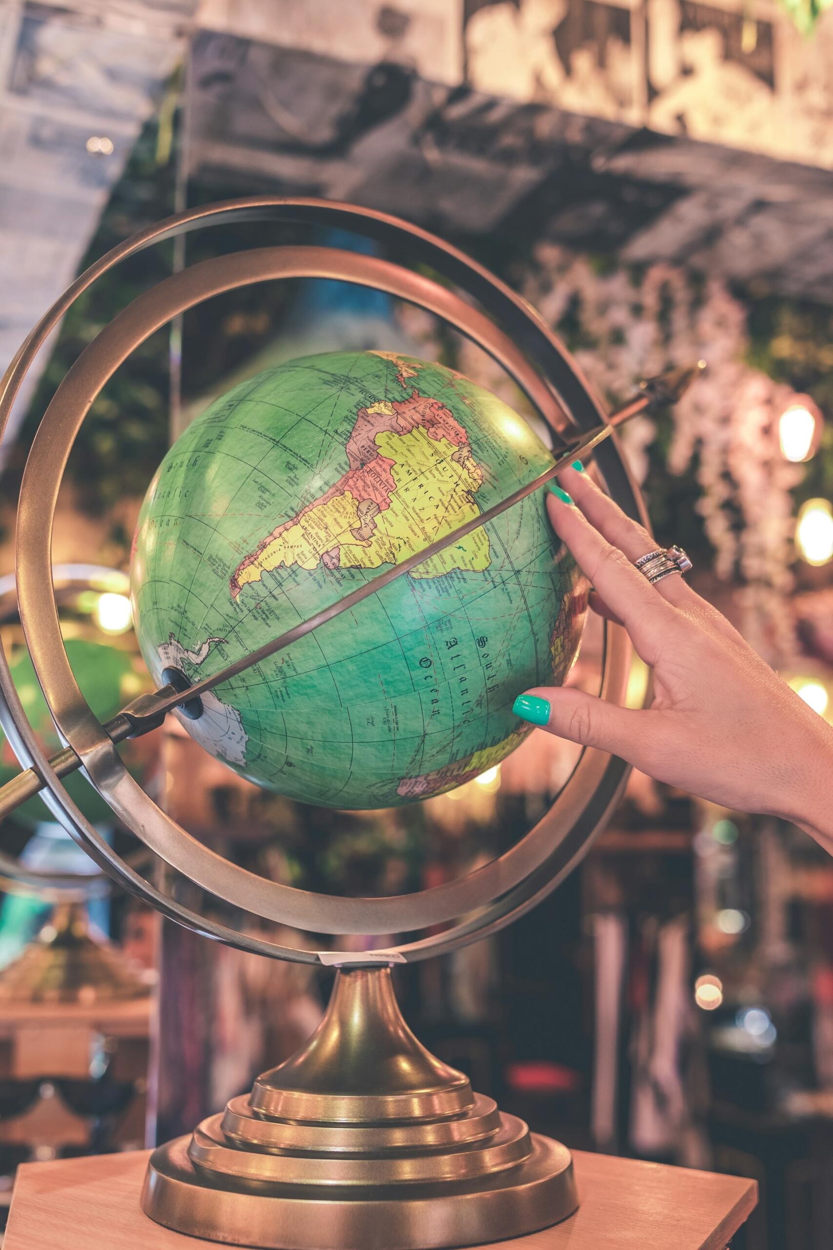 A hand with teal nail polish gently spins a globe on a golden stand, surrounded by a cozy, softly lit background.