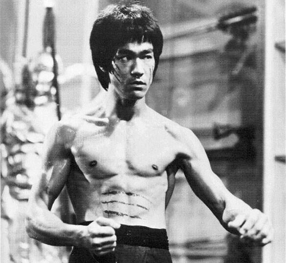 A muscular Bruce Lee stands in a fighting stance, showcasing defined abs and notable scars on their torso, background softly blurred.