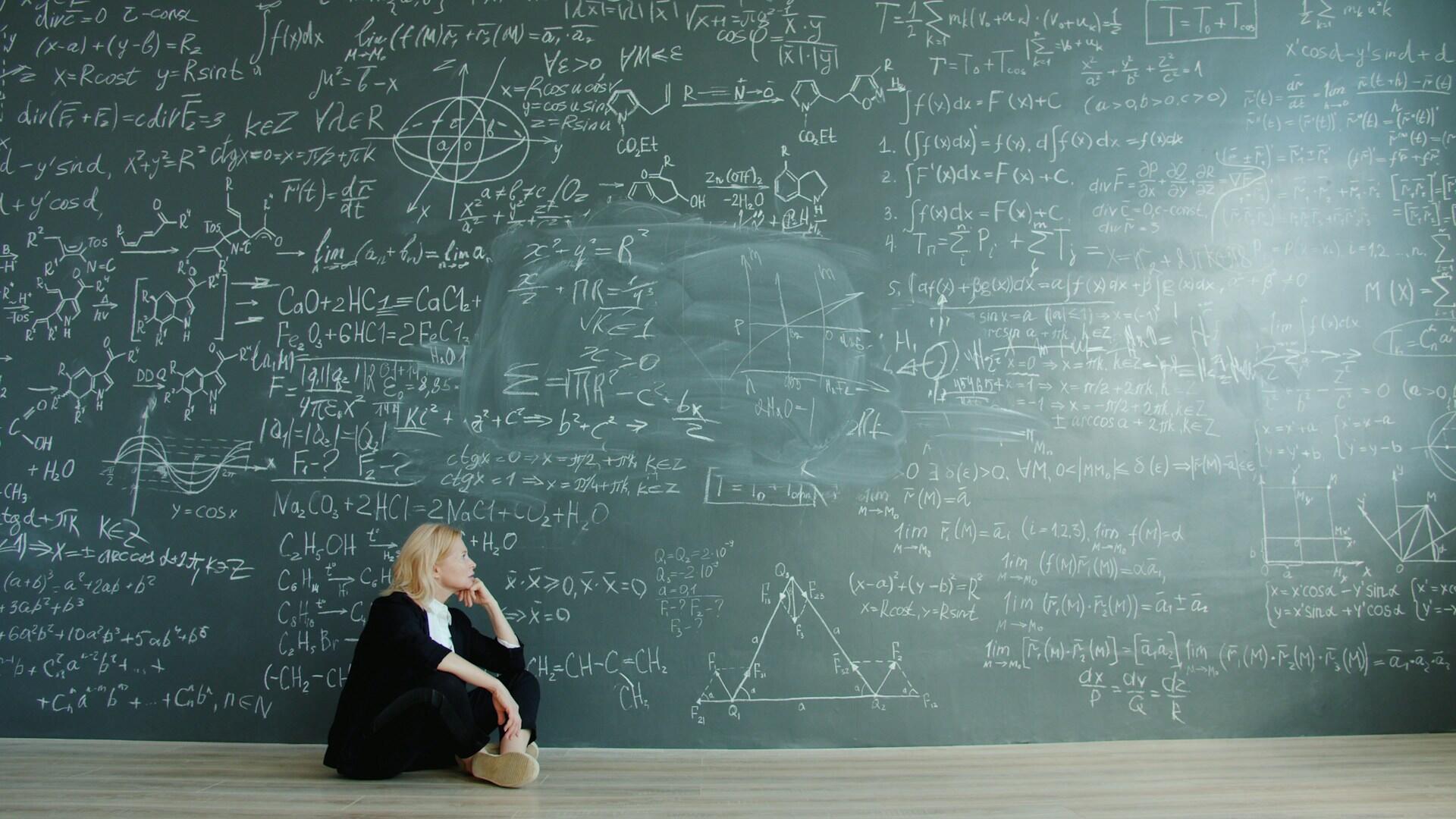 A person sits in front of a chalkboard filled with intricate mathematical equations and scientific diagrams, reflecting deep contemplation.