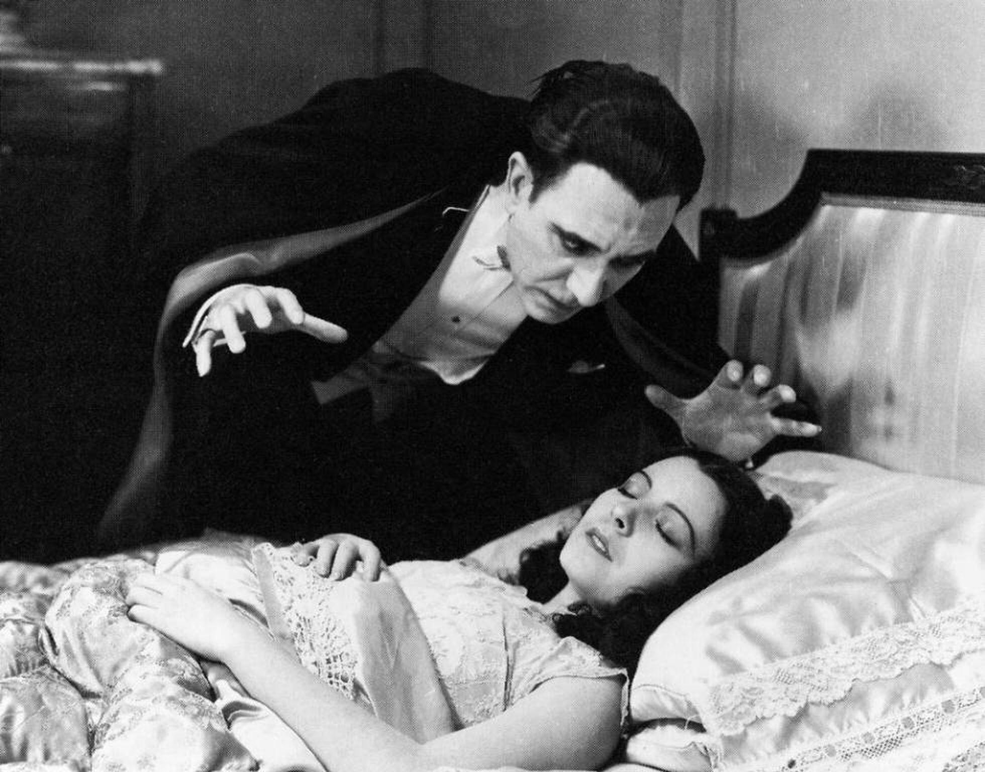A man in a vintage tuxedo and cape leans over a woman lying on a bed, creating a dramatic, suspenseful scene.