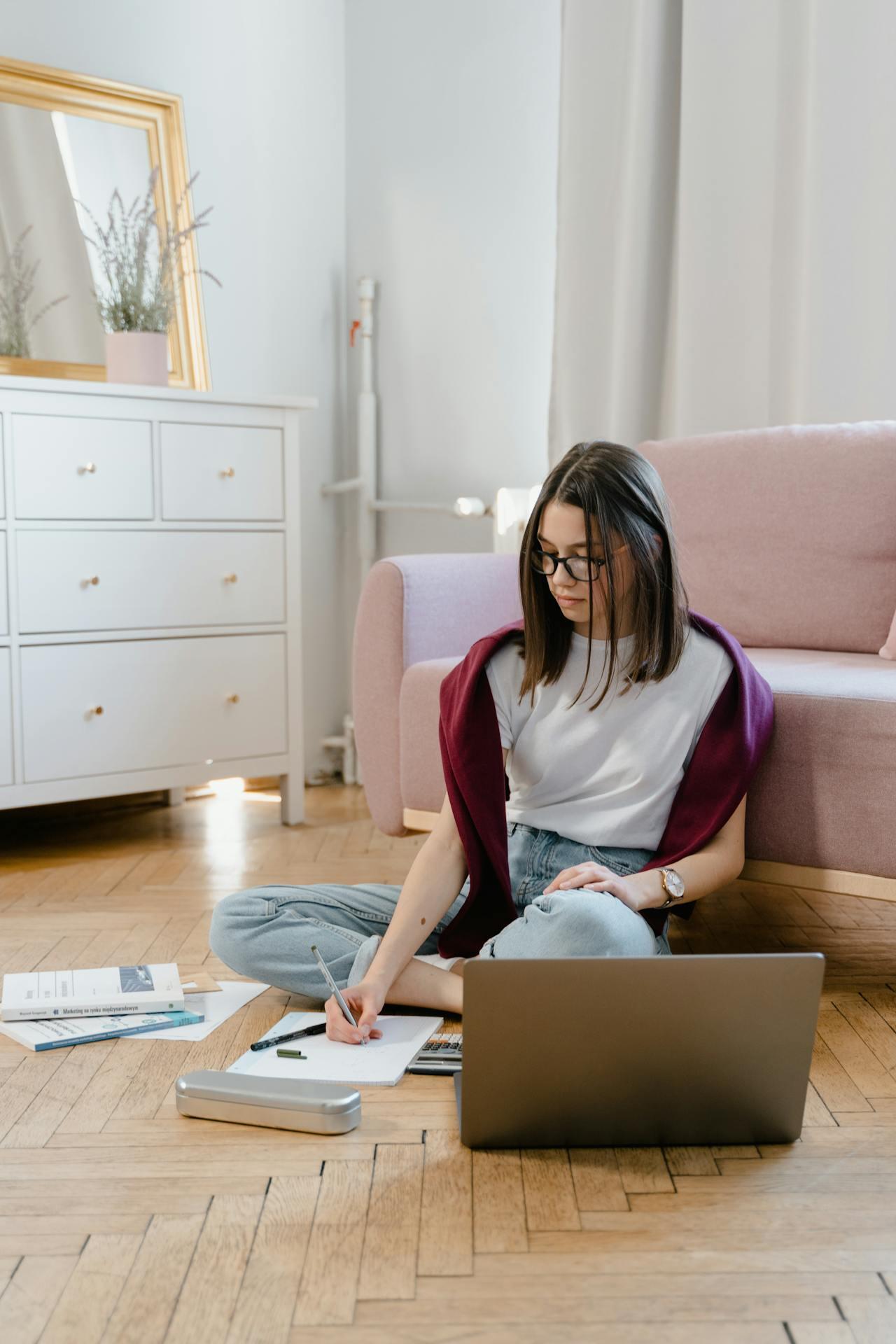 A person sitting cross-legged on the floor, working on a laptop surrounded by papers and a pencil, in a cozy living space.