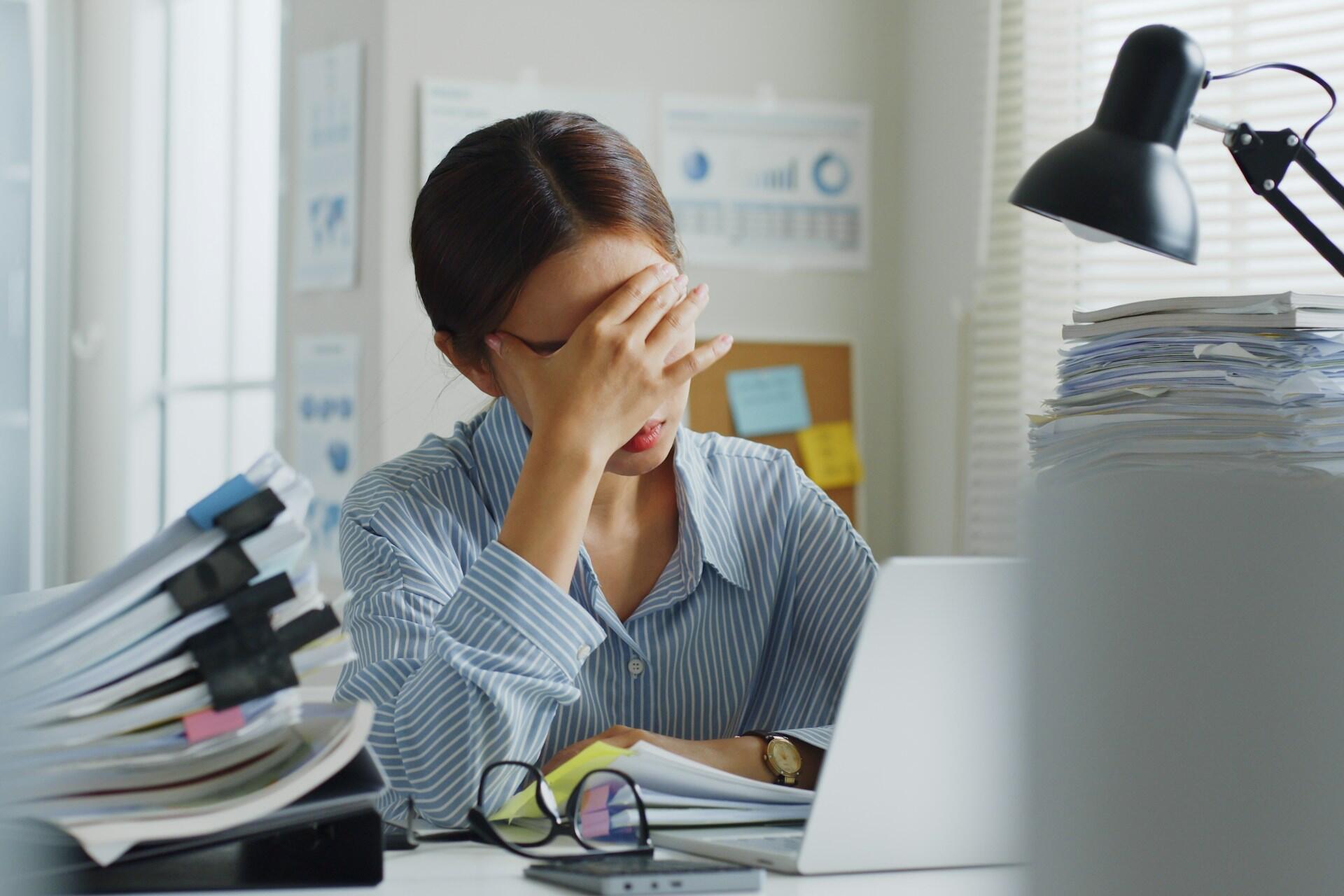 A person holds their head in their hand sitting next to a pile of paperwork.