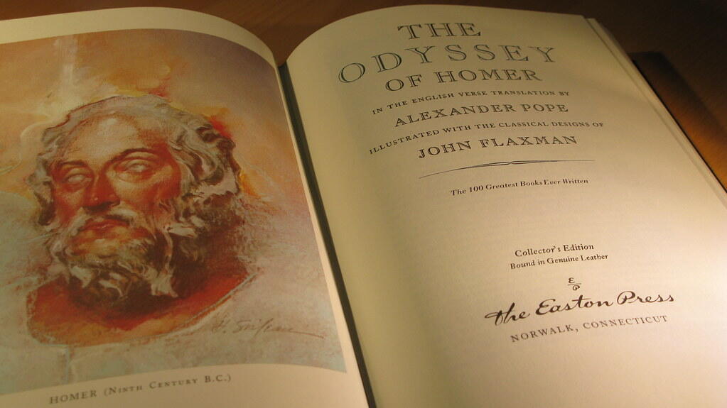 An open copy of "The Odyssey of Homer," featuring elegant typography and classical illustrations, resting on a wooden surface.
