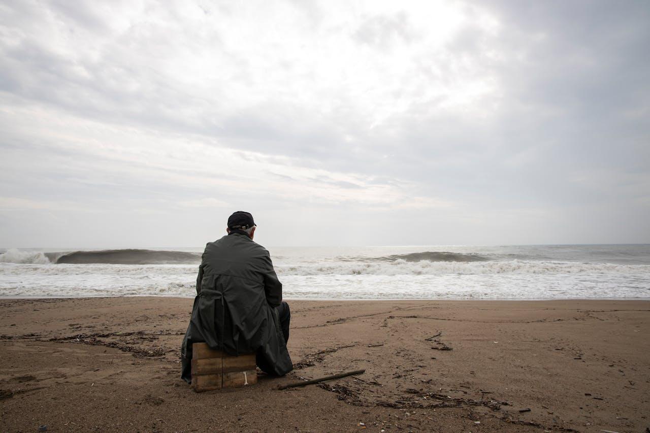 A man in a dark coat sits on a wooden box facing the ocean, watching waves under a cloudy sky at the beach.