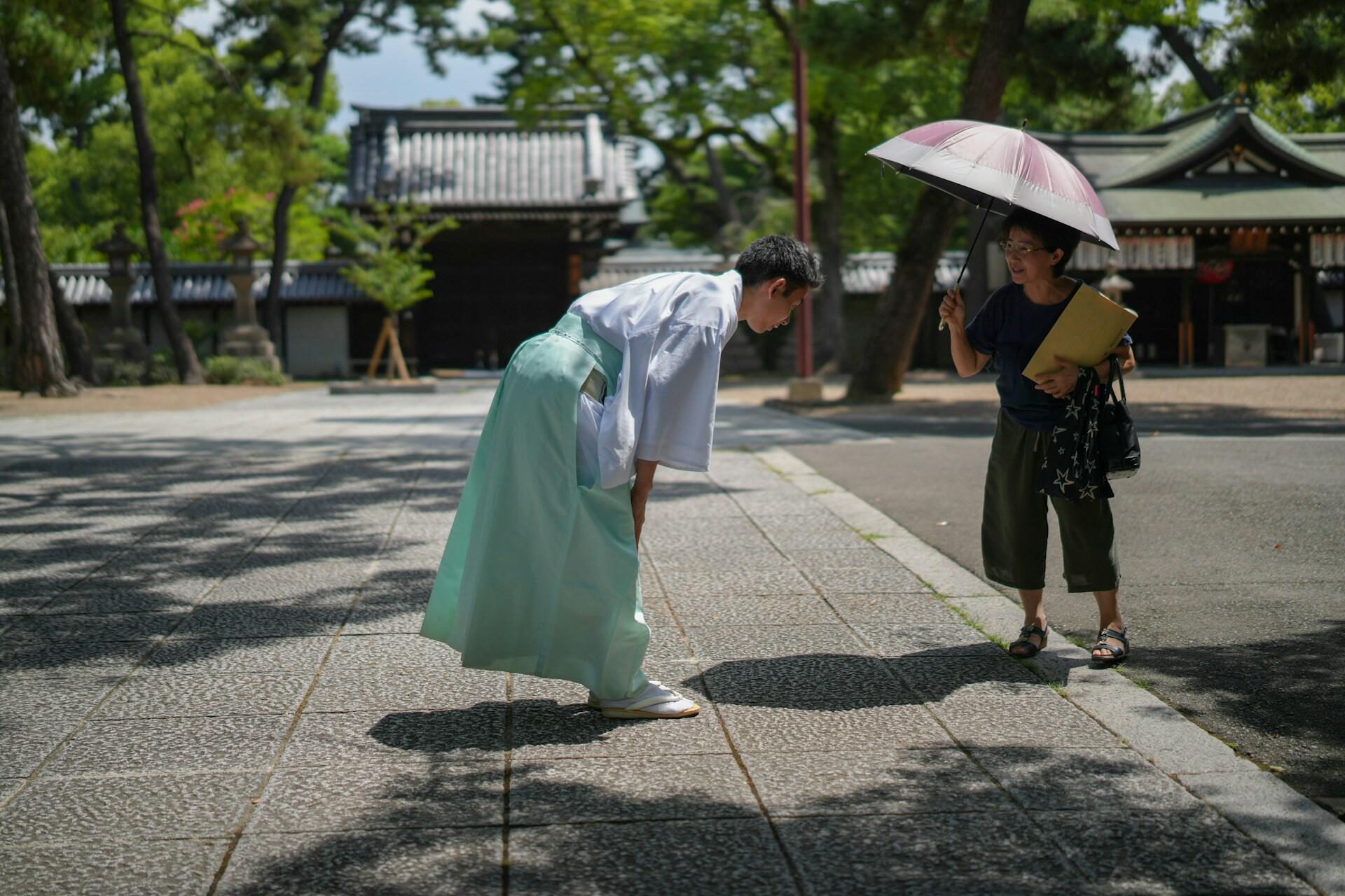 A person in traditional attire bows respectfully while a woman under a pink umbrella holds a folder, set in a serene outdoor environment.