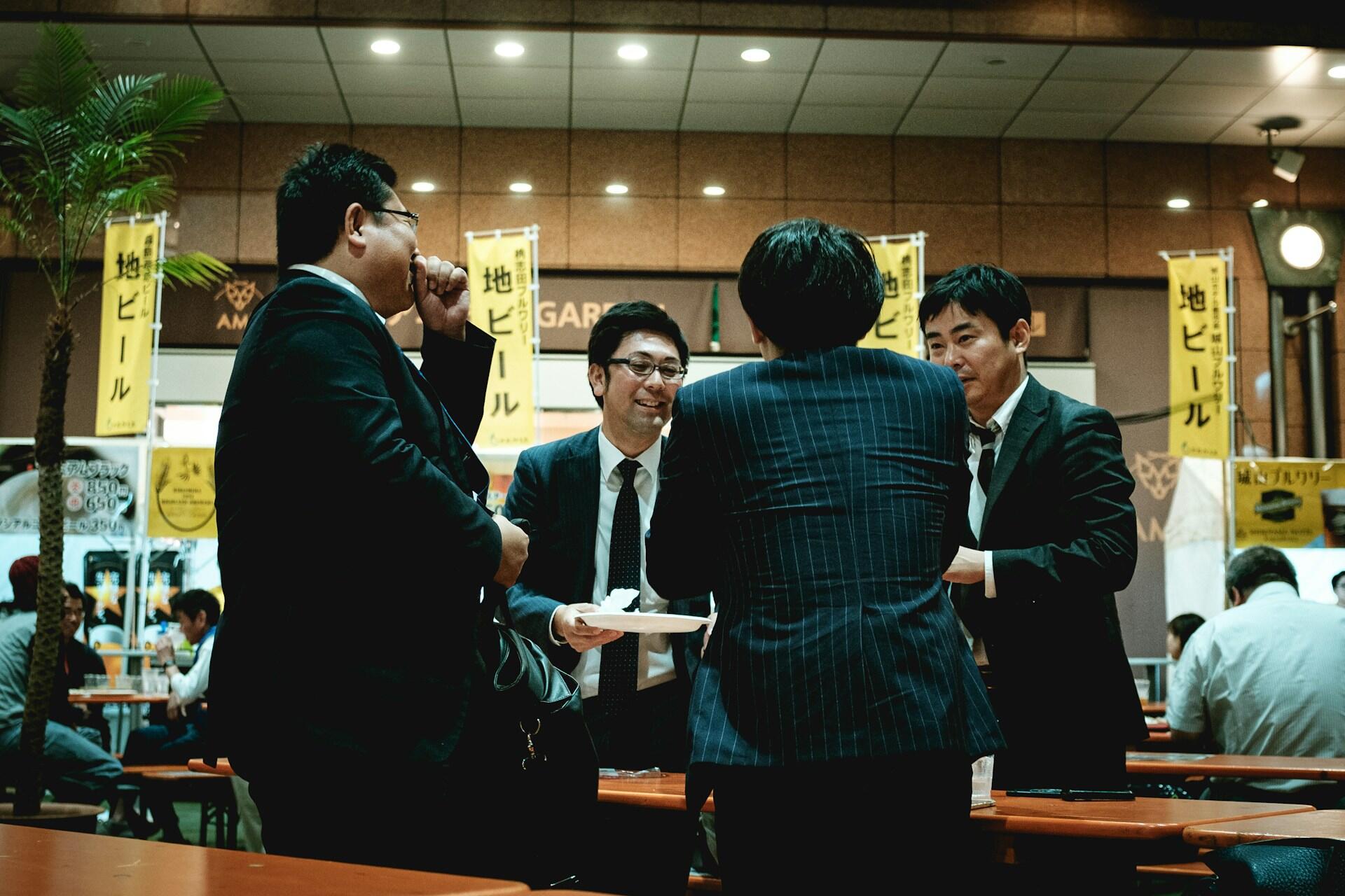 A group of business professionals in suits engage in conversation at a busy indoor venue with promotional banners in the background.