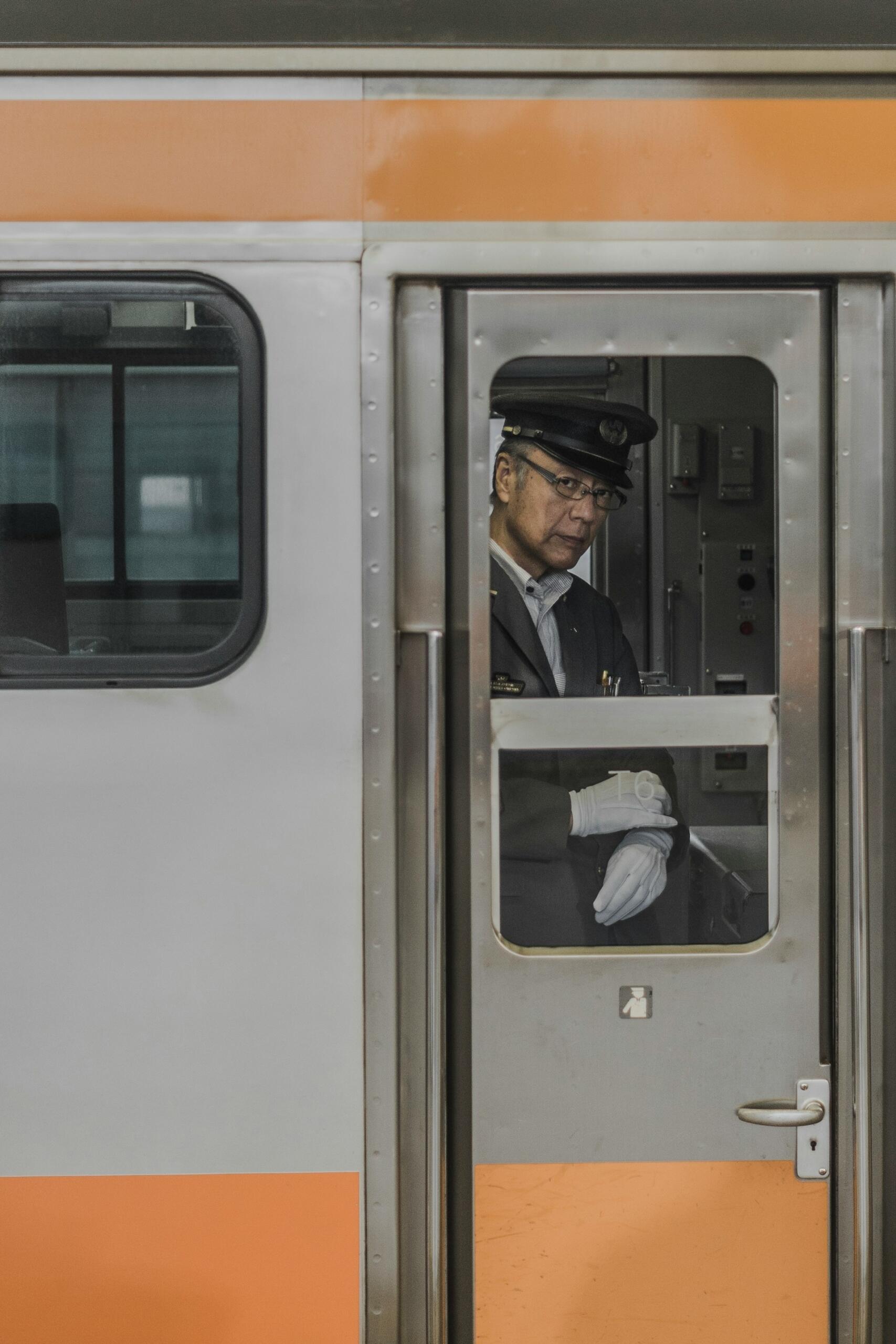 A train conductor in a uniform stands in the doorway of a subway car, wearing white gloves and a cap, with machinery in the background.