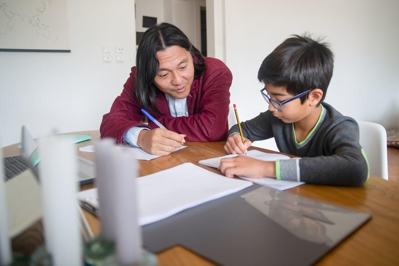 A woman and a child engage in a focused writing activity at a wooden table, with notebooks and a laptop present.