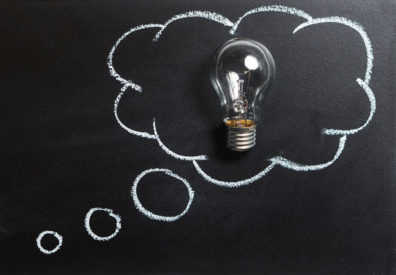 A light bulb is surrounded by a chalk-drawn thought bubble on a black chalkboard, symbolizing ideas and inspiration.