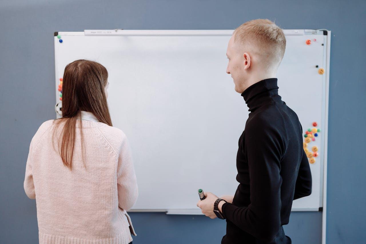 Two individuals stand in front of a blank whiteboard, one holding a marker, while colorful magnets decorate the board's edges.