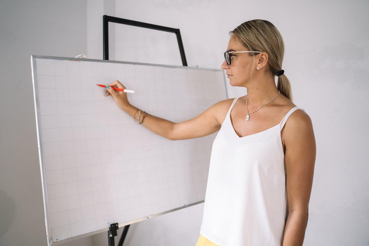 A woman in a white tank top is writing on a blank whiteboard with a red pen, emphasizing a collaborative or instructional setting.
