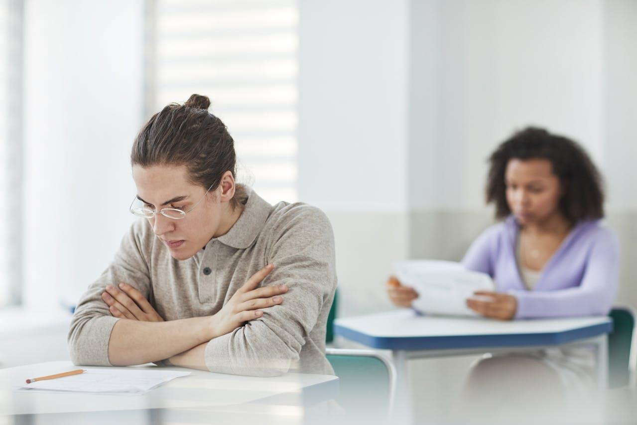Two students sit at desks in a bright classroom, focusing intently on their papers amidst a quiet testing atmosphere.