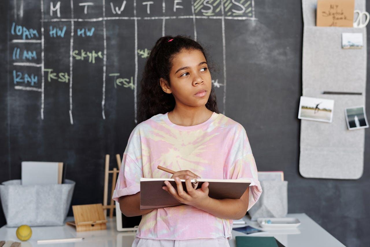 A young person in a colorful tie-dye shirt holds a notebook and pencil, standing in front of a chalkboard with a weekly schedule.