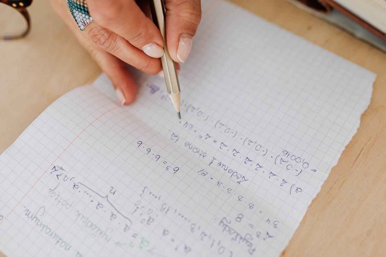 A hand holding a pencil writes complex mathematical equations in a grid notebook on a wooden surface.