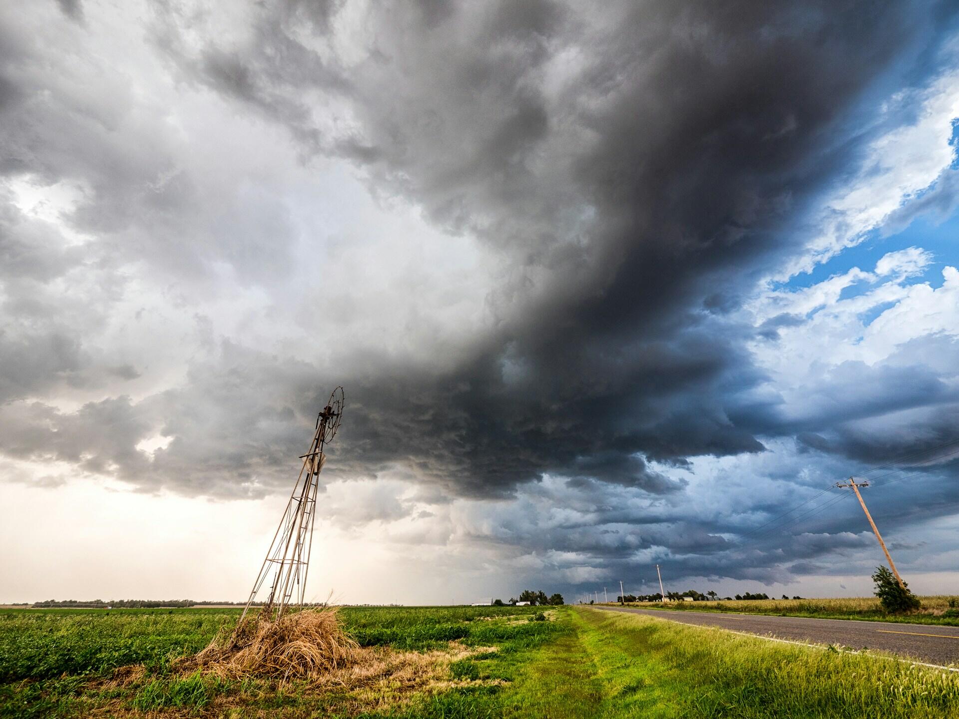 A weathered windmill stands in a field beneath a dramatic sky filled with dark storm clouds and streaks of blue.