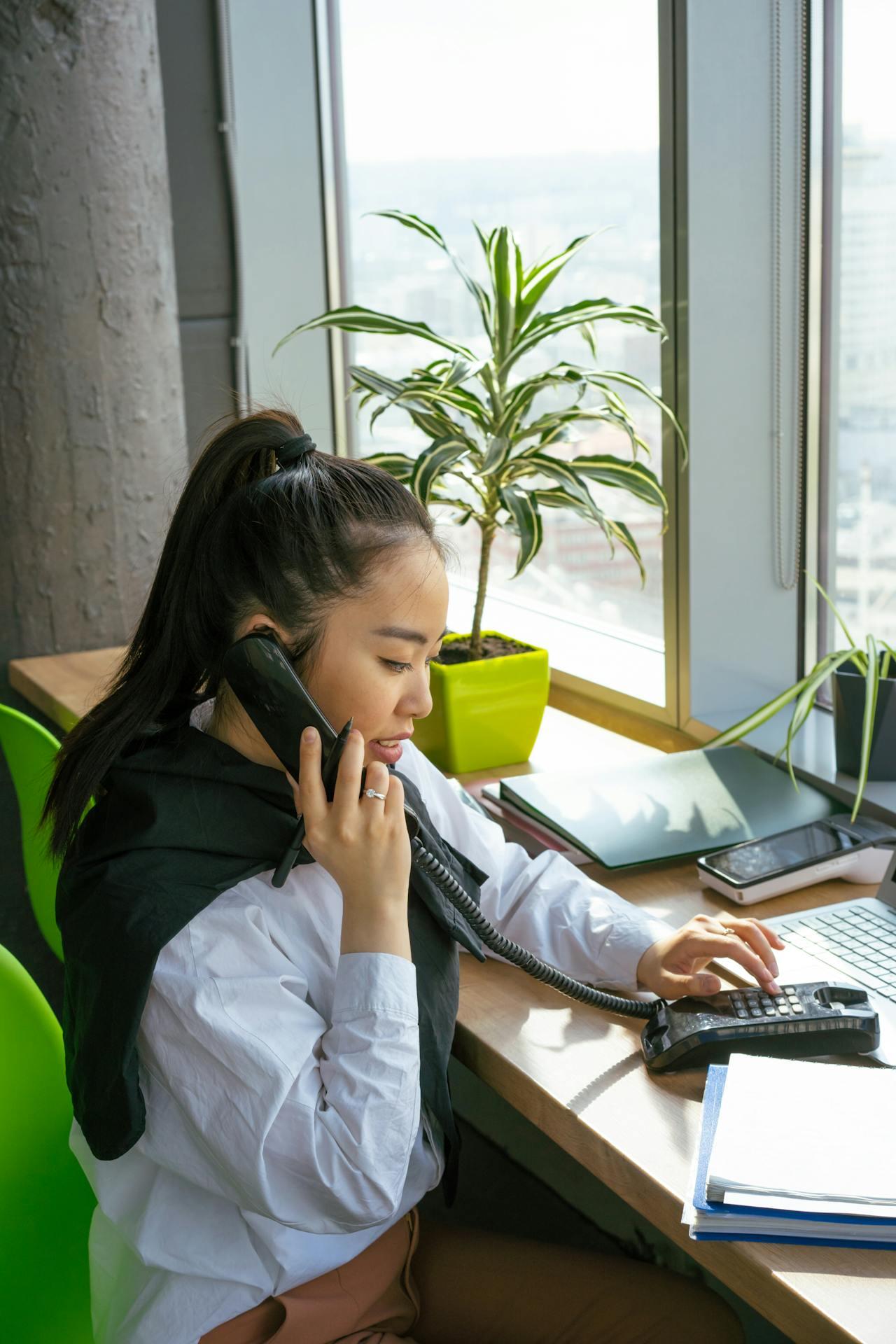 A person sits at a desk with a phone, laptop, and documents, while a green plant sits on the windowsill in a bright office setting.