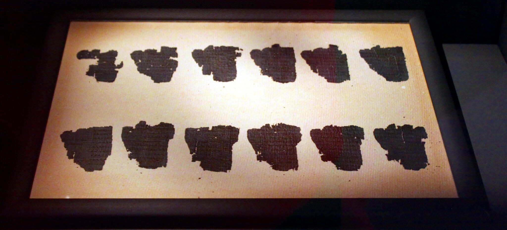 Ancient fragments displayed in a frame, featuring irregular black shapes resembling pieces of parchment or scrolls on light background.