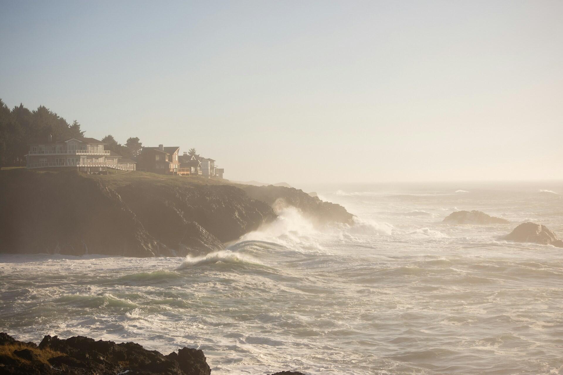 Coastal scene with waves crashing against rocky cliffs, framed by misty light and distant oceanfront homes under a pale sky.