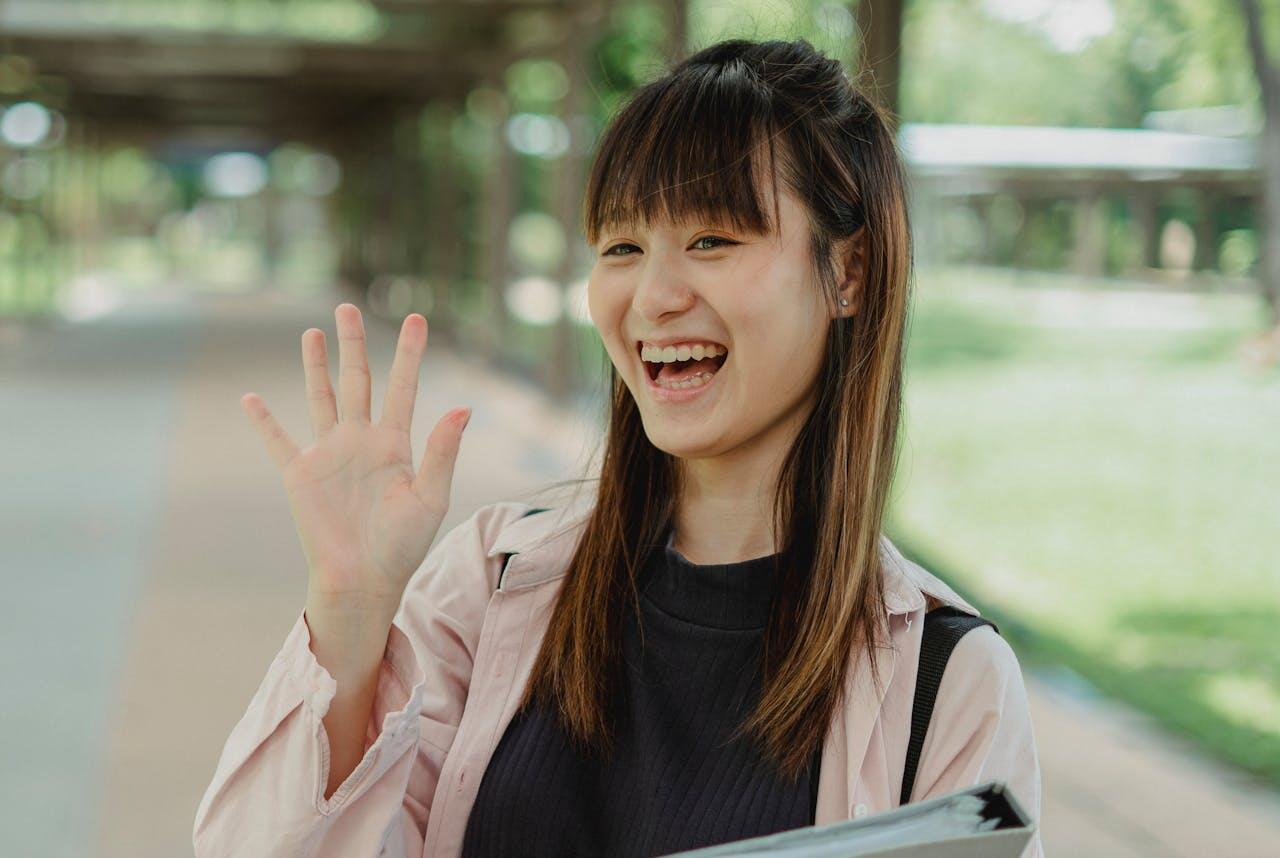 A young woman in a light pink jacket waves her hand while standing in a green outdoor corridor, holding a folder.